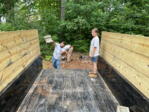 A view from the back of a pick up truck. A man can be seen tossing a cinder block into the left side of the truck, while a woman stands on the right.