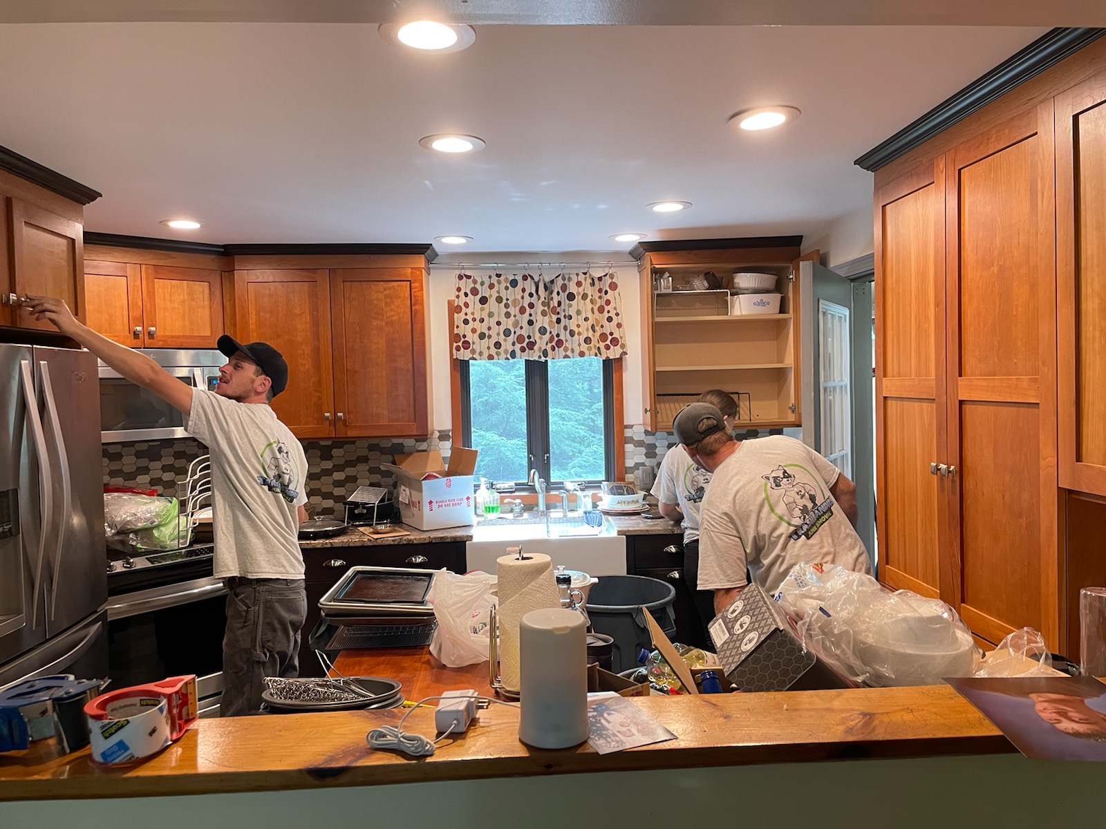 A view of a kitchen. Many drawers align the top of the room, a fridge is present on the left side, and cabinets on the right. Two workers clean the area and move things around.