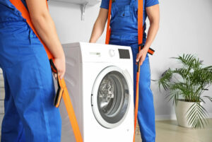 Two movers in all blue uniforms use orange belts to remove an old washing machine from a white room.