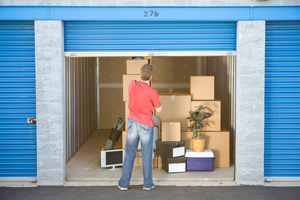 A man opens a blue garage door to reveal a storage unit with several stacks of boxes inside.