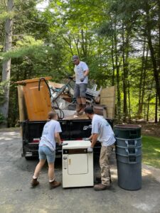 Three workers load a pickup truck with junk items. You can see several appliances and trash bins among the rubble. A forest of trees lies in the background.