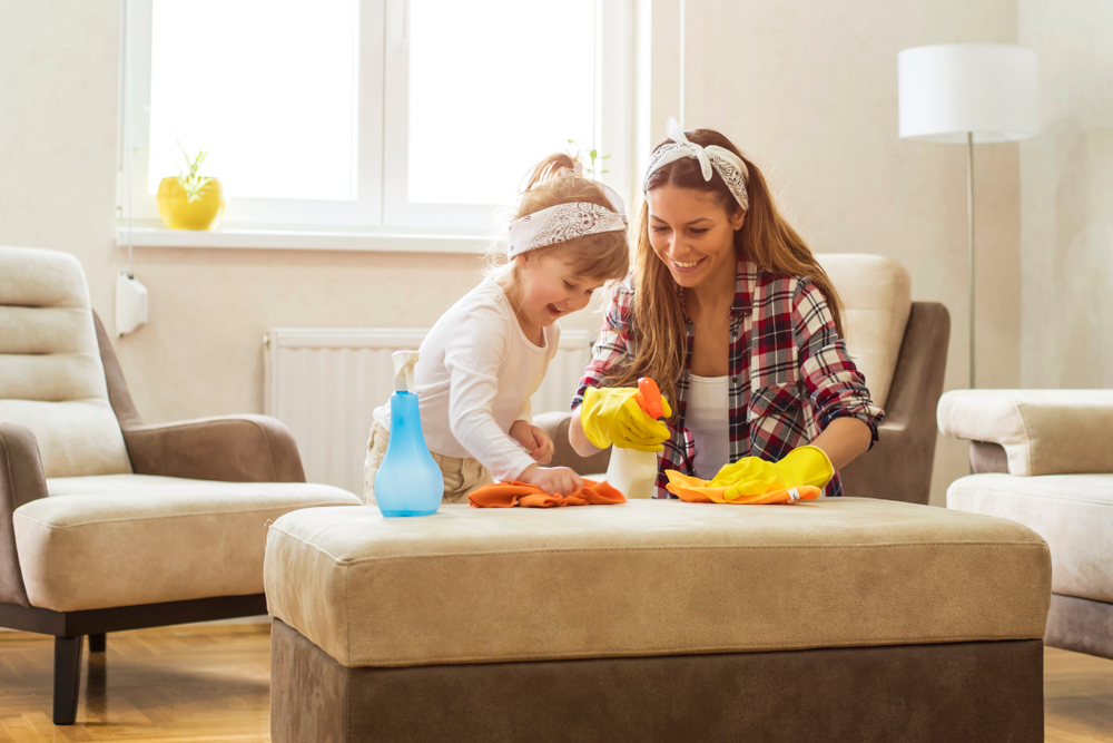 A smiling mother and young daughter use an orange rag to clean the top of a couch cushion. You can see the blue spray bottle on top of the cushion and the yellow gloves worn by the mother as well.