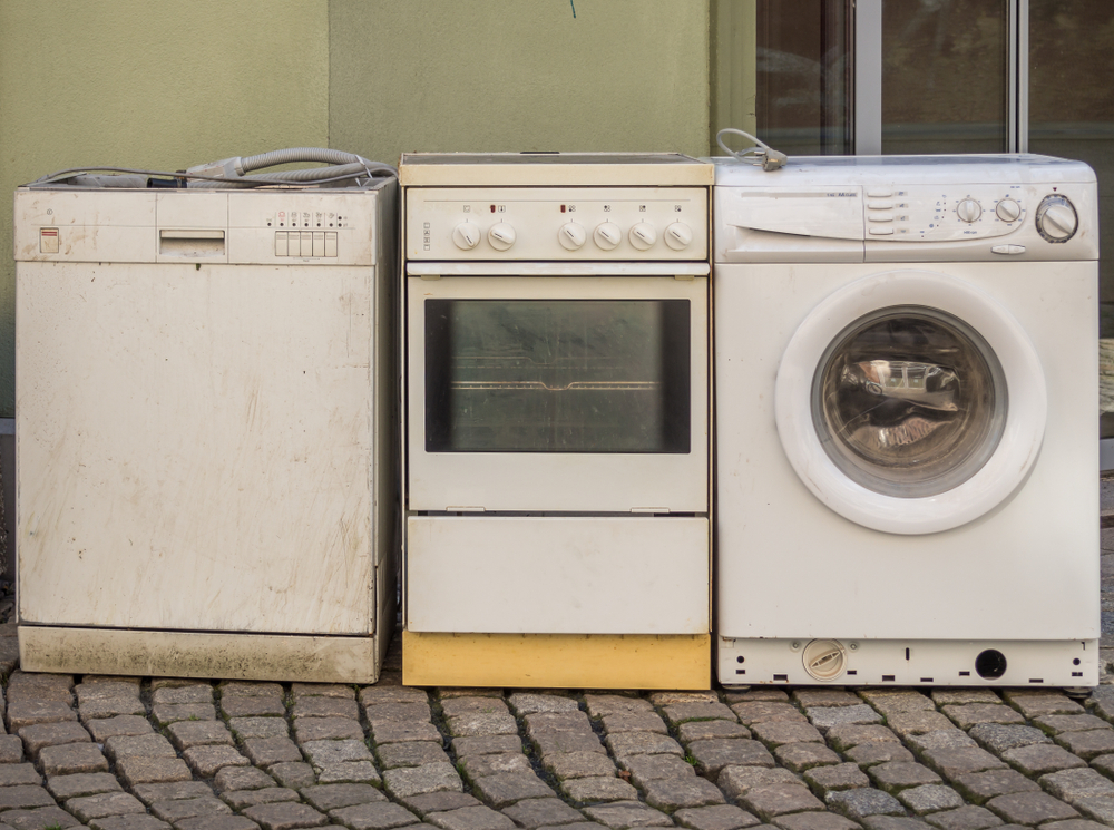 An old dishwasher, oven, and washer sit next to each other on a concrete brick road.