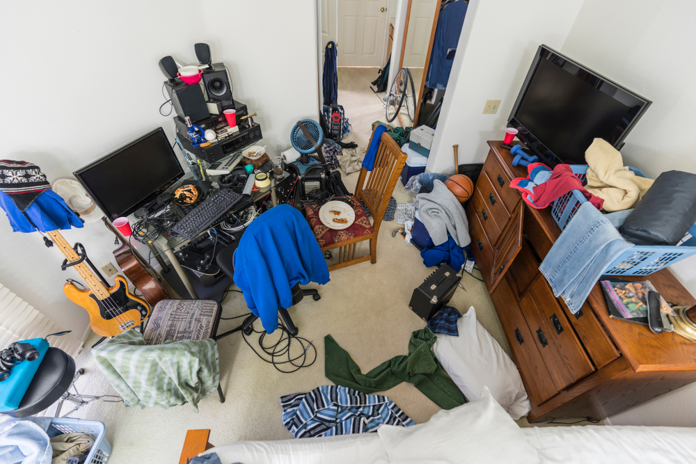 An image of a bedroom filled with clutter. You can see various pieces of furniture, clothing, and trash littered amongst the clutter.